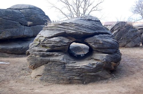 A large, weathered rock with a circular hole in the center, surrounded by other similar rocks and bare trees.