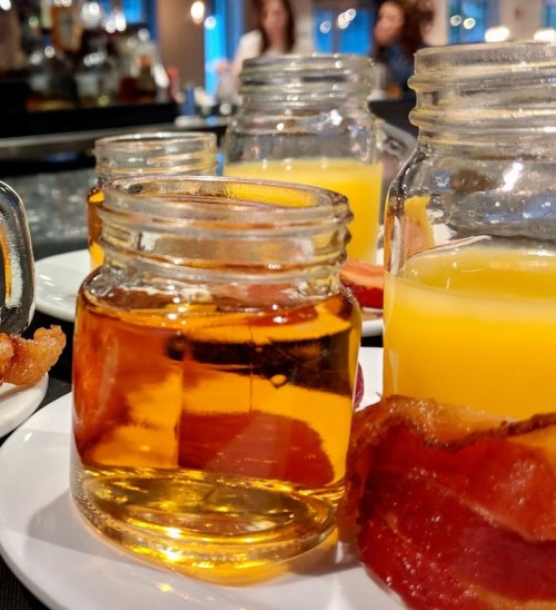 A close-up of jars filled with amber liquid and orange juice, accompanied by crispy bacon on a plate.