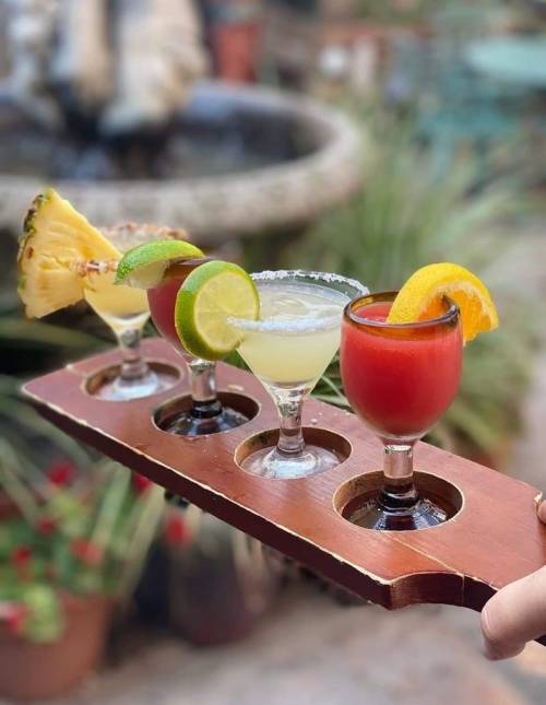 A wooden tray holds four colorful cocktails, garnished with fruit, in a vibrant outdoor setting.