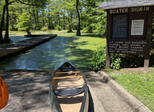A canoe at a boat launch area with a sign about camping permits and regulations in a green, wooded setting.