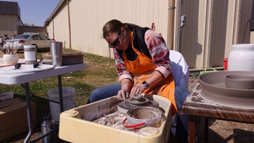A person in an orange apron shapes clay on a pottery wheel outdoors, surrounded by tools and pottery materials.