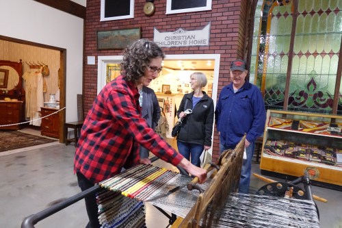 A woman in a red plaid shirt weaves at a loom, while two people observe in a museum setting.