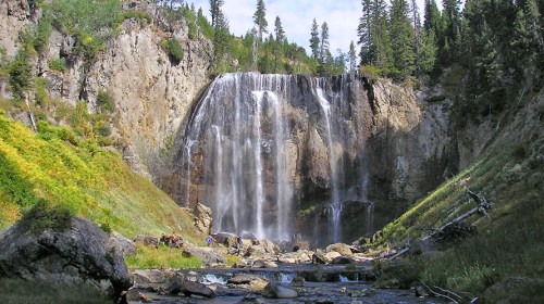 A stunning waterfall cascades down rocky cliffs, surrounded by lush greenery and tall trees under a bright blue sky.