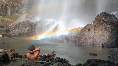 A person relaxes by a waterfall, with a rainbow arching over the rocks and water in a serene natural setting.