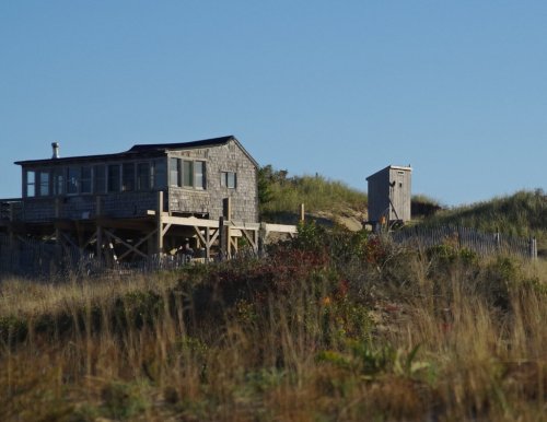 A rustic beach house on stilts, surrounded by grass and sand dunes, with a small shed nearby under a clear blue sky.