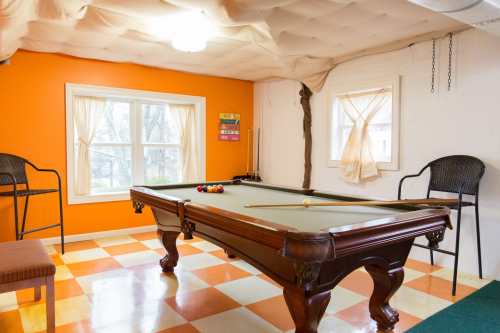 A bright game room featuring a pool table, orange walls, and two windows with sheer curtains.