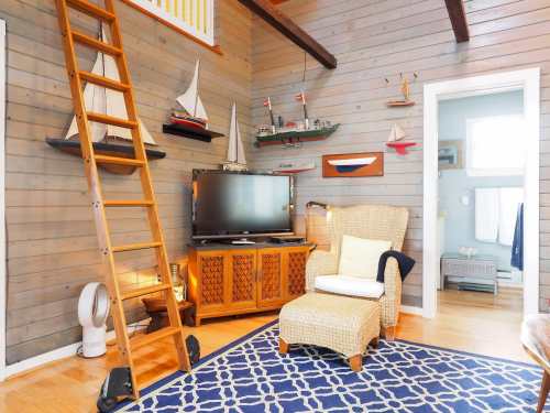 Cozy living room with a ladder, TV, wicker chair, and nautical decor on wooden walls. Blue patterned rug on the floor.
