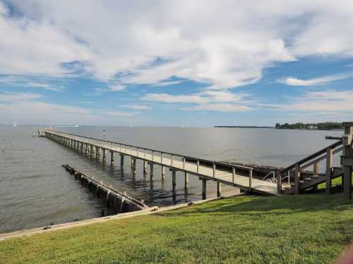 A long wooden pier extends into calm waters under a blue sky with scattered clouds. Green grass lines the shore.