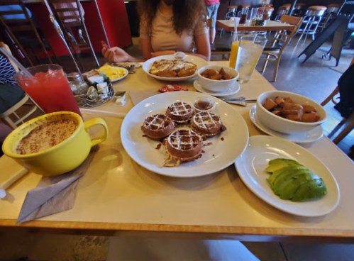 A table filled with various breakfast dishes, including waffles, avocado, and drinks, with a person partially visible.