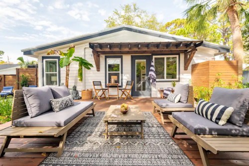 Cozy outdoor patio with seating, a table, and tropical plants, in front of a charming white house.