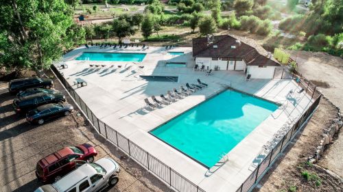 Aerial view of a pool area with two swimming pools, lounge chairs, and a nearby parking lot. Green trees in the background.