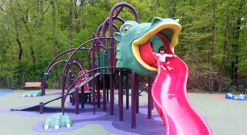 A child slides down a bright red slide shaped like a dragon's mouth at a colorful playground surrounded by trees.