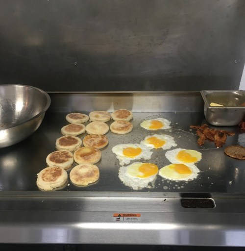 A griddle with cooking eggs, English muffins, and bacon, alongside a mixing bowl and a small container.