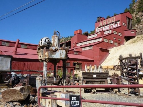 Historic Argo Gold Mine and Mill with red buildings and mining equipment in the foreground, set against a clear blue sky.