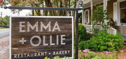 Sign for "Emma + Ollie Restaurant + Bakery" in front of a charming building with greenery and outdoor seating.