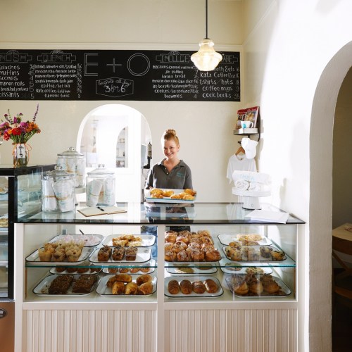 A bakery display case filled with various pastries, with a smiling employee behind the counter and a chalkboard menu above.