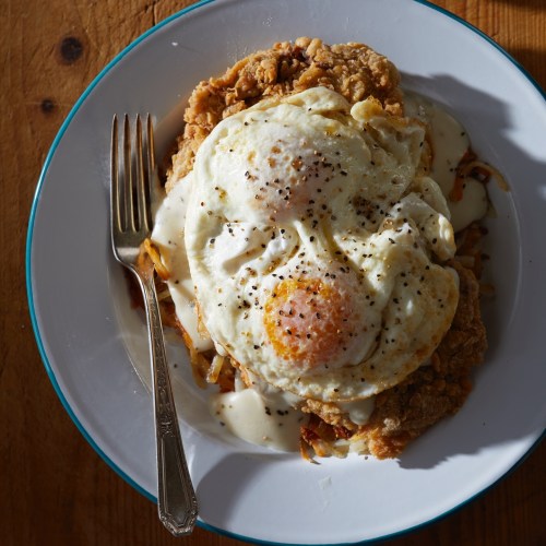 A plate of fried chicken topped with two sunny-side-up eggs and creamy gravy, served with a fork on the side.