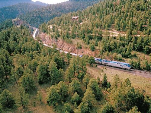 A train travels along a winding track through a lush, green forested landscape with mountains in the background.