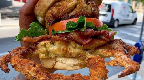 A hand holding a large sandwich with crab, bacon, lettuce, and tomato, set against an outdoor background.