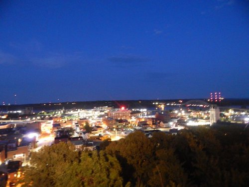 A nighttime cityscape with illuminated buildings and a dark blue sky, surrounded by trees and distant lights.