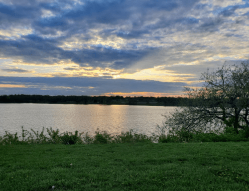 A serene lake at sunset, with colorful clouds reflecting on the water and greenery in the foreground.
