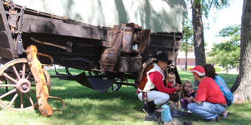 A person in historical attire teaches children by a campfire near a covered wagon in a grassy area.