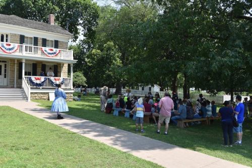 A gathering of people outside a historic building, with a woman in a blue dress and patriotic decorations.