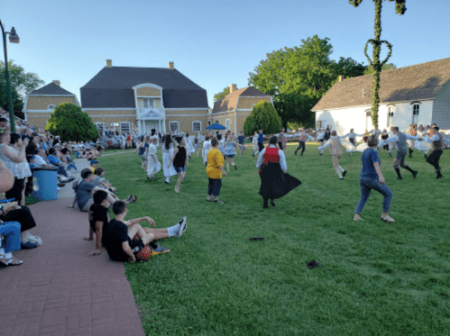 A lively outdoor dance performance with a crowd watching on a grassy area, surrounded by historic buildings.