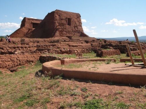 Ancient adobe ruins on a grassy landscape under a clear blue sky, with a circular structure in the foreground.