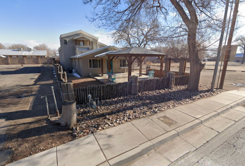 A two-story house with a porch, surrounded by a gravel yard and trees, viewed from the street.