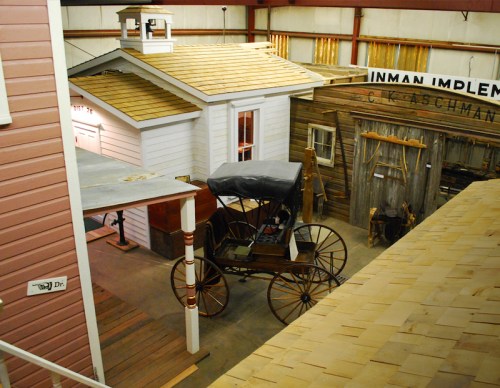 A vintage horse-drawn carriage in a museum setting with replica buildings and a wooden structure in the background.