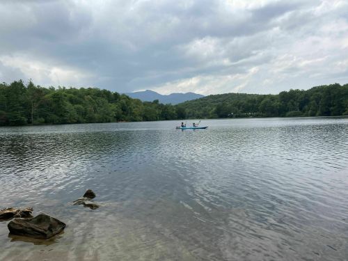 A calm lake surrounded by trees, with two people kayaking in the distance under a cloudy sky.
