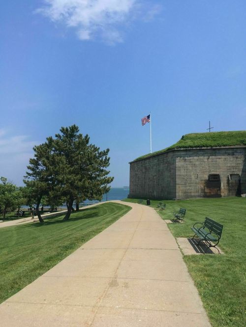 A pathway leads to a stone fort with a flag, surrounded by green grass and trees under a clear blue sky.