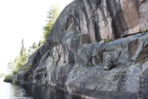 A rocky cliff rises above calm water, surrounded by trees and a clear sky.
