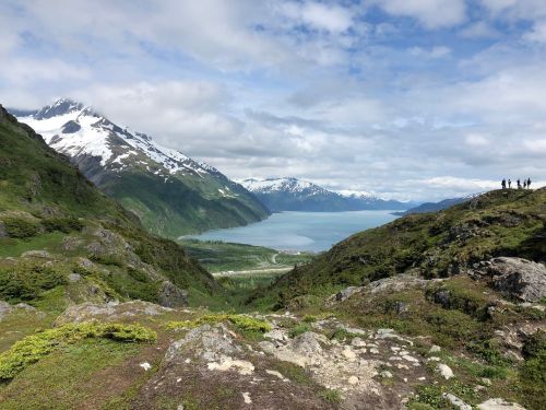A scenic view of mountains and a lake under a partly cloudy sky, with hikers visible in the distance.