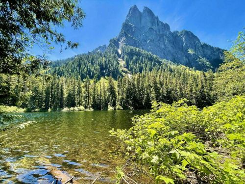 A serene lake surrounded by lush green trees, with a towering mountain in the background under a clear blue sky.