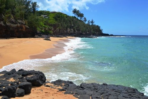 A serene beach with golden sand, gentle waves, and lush greenery along the rocky coastline under a clear blue sky.