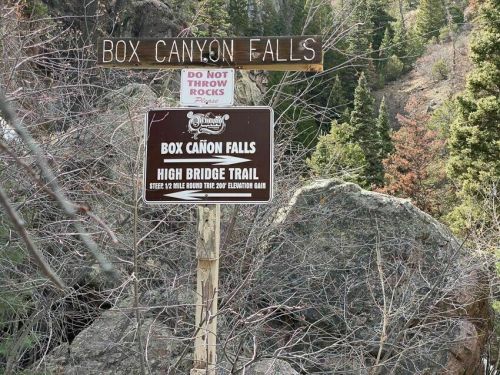Sign for Box Canyon Falls with trail information and a warning against throwing rocks, surrounded by trees and rocks.