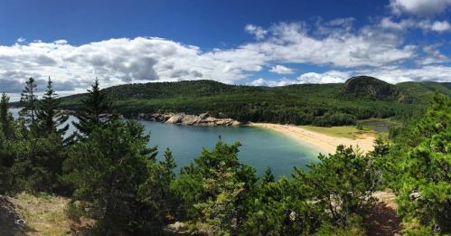 A scenic view of a sandy beach surrounded by lush green hills and a calm blue water under a partly cloudy sky.