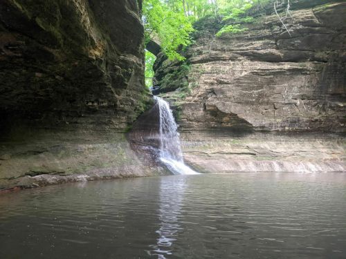 A serene waterfall cascades into a calm pool, surrounded by rocky cliffs and lush green foliage.