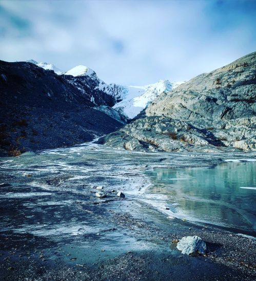 A serene landscape featuring a glacier, rocky terrain, and a calm body of water under a cloudy sky.
