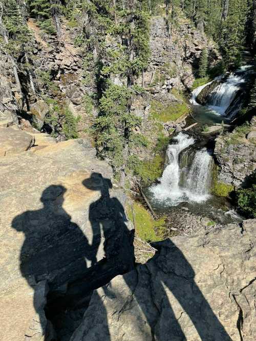 Silhouettes of two people holding hands on a rocky ledge overlooking a waterfall and lush greenery.