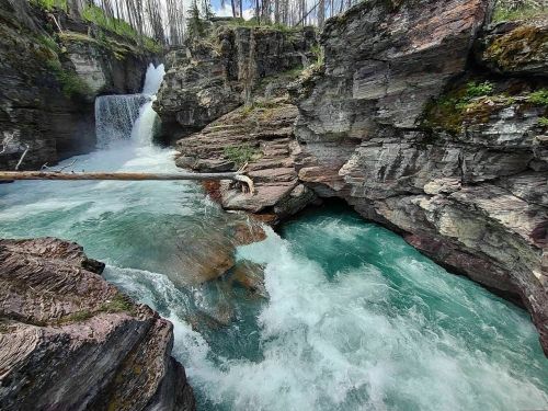 A vibrant turquoise river flows through rocky cliffs, with a waterfall cascading in the background amidst lush greenery.