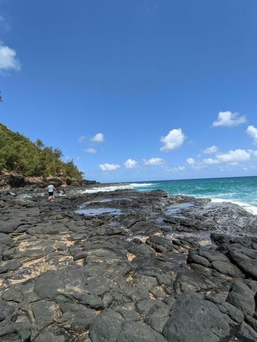 A rocky shoreline with a person walking along the coast, under a clear blue sky and fluffy white clouds.