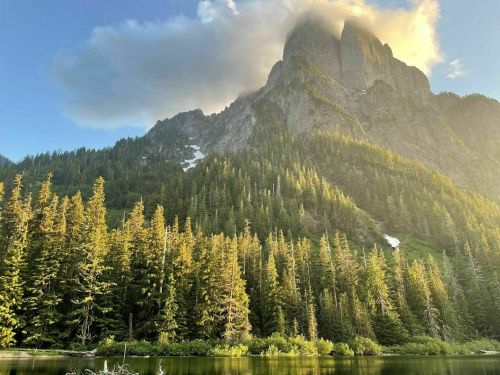 Majestic mountains rise above a serene lake, surrounded by lush green forests and soft clouds in the sky.