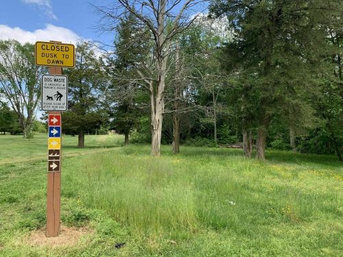 Sign indicating park hours (closed dusk to dawn) and dog waste disposal, surrounded by trees and tall grass.