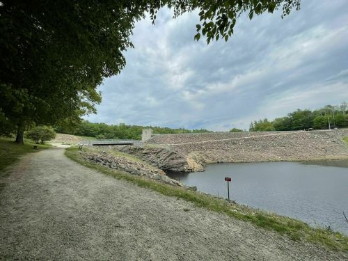A gravel path alongside a calm lake, with a dam and trees under a cloudy sky in the background.