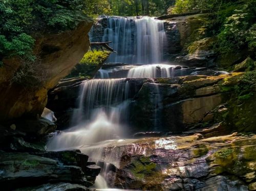 A serene waterfall cascading over rocky ledges, surrounded by lush green foliage and sunlight filtering through the trees.