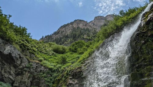 A cascading waterfall flows down rocky cliffs, surrounded by lush greenery and mountains under a blue sky.
