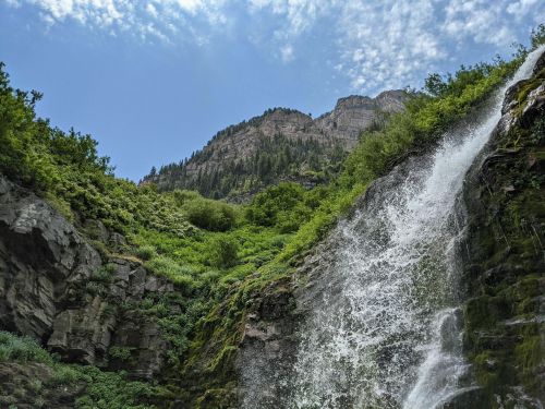 A cascading waterfall flows down rocky cliffs, surrounded by lush greenery and a blue sky with scattered clouds.